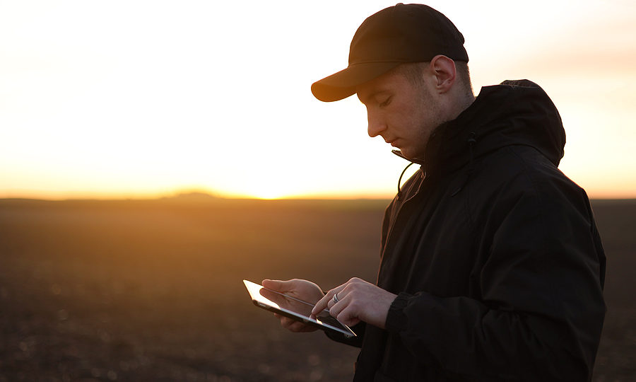 Farmer uses digital tablet on field with plowed soil at sunset to look over his farm bookkeping.