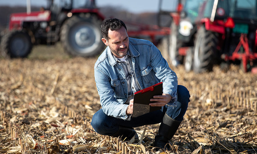 Mature adult farmer squatting in field, writing notes on notepad about his farm budget while tractors working in background.